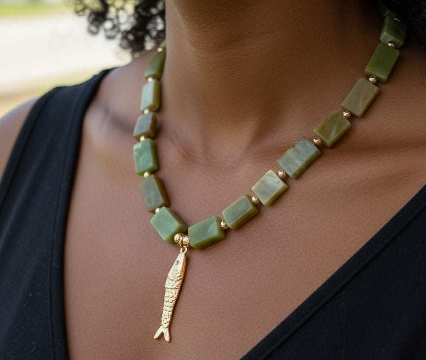 A beaded necklace with olive green rectangular beads and a small orange fish charm, displayed on a pink jewelry bust at an outdoor market.