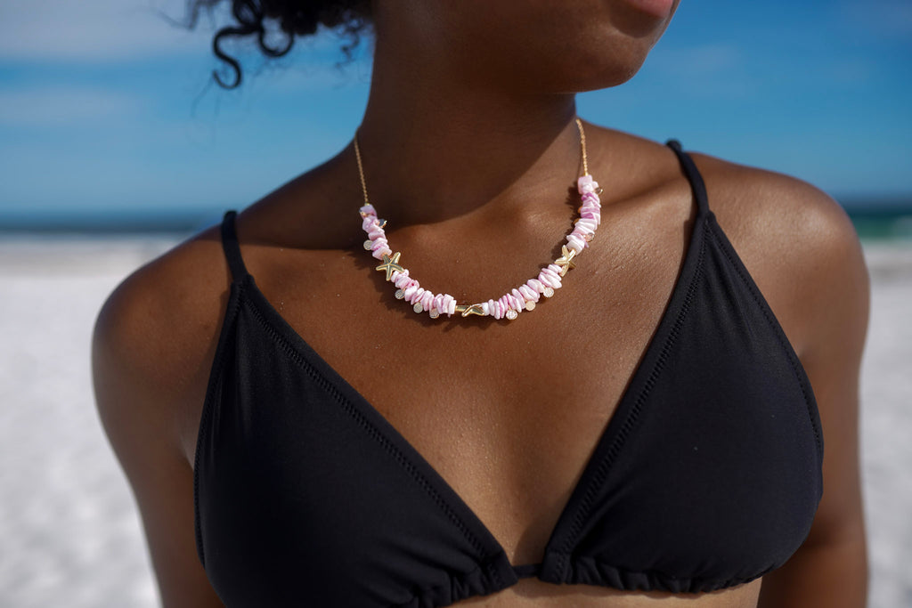 A close-up of a woman on a beach wearing a shell necklace with gold-toned chain links. The gillian necklace features light pink and white seashells with small gold accents, worn over a black bikini top against a blue sky and ocean background.