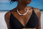 A close-up of a woman on a beach wearing a shell necklace with gold-toned chain links. The gillian necklace features light pink and white seashells with small gold accents, worn over a black bikini top against a blue sky and ocean background.