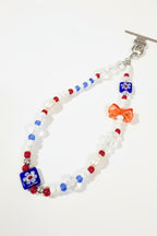 A beaded bracelet with a silver toggle clasp, featuring white faux-pearl beads, red and blue accent beads, clear faceted beads, and a small red bow charm