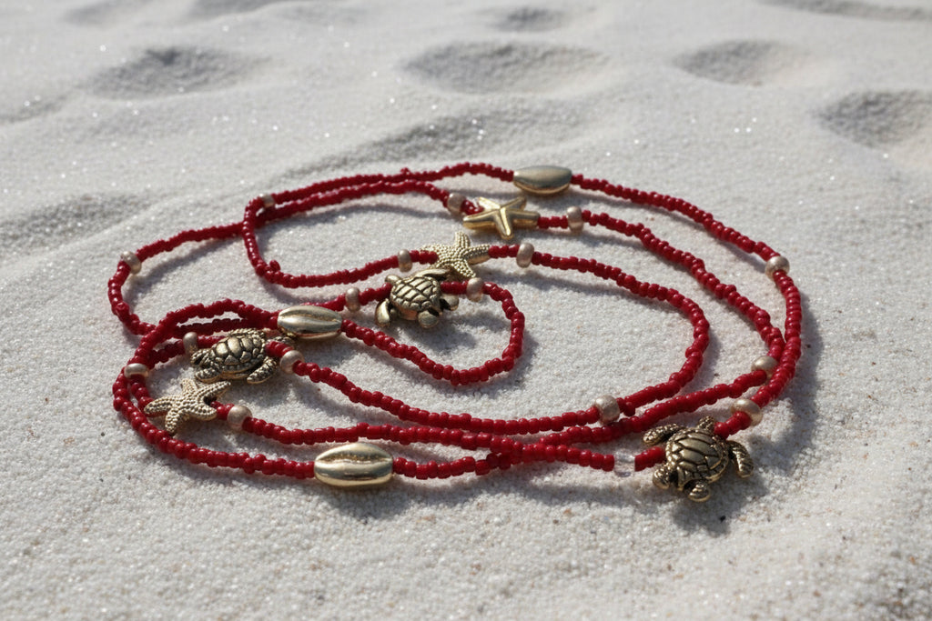 Red waistbeads with gold turtle and starfish charms on a white sandbackground